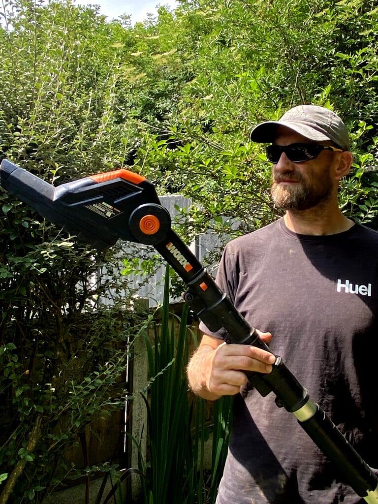 Gardener holding the Worx WG252E cordless pole hedge trimmer with extended shaft in a sunlit garden